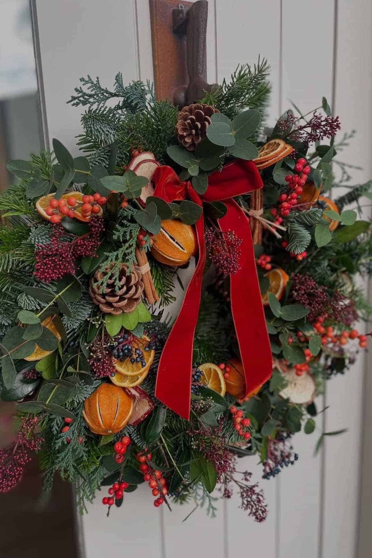 A festive wreath with pine branches, pinecones, dried orange slices, red berries, and a red velvet bow hangs on a door.