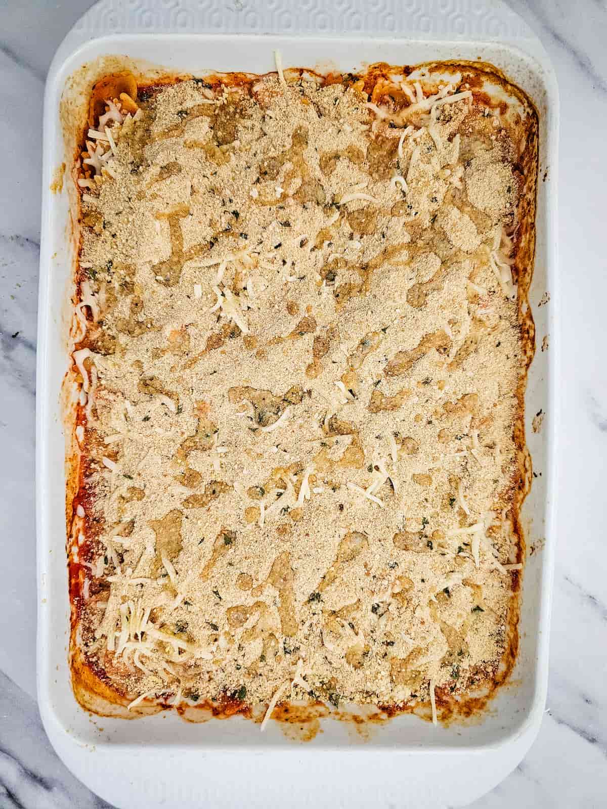 A rectangular white baking dish filled with pasta topped with a layer of breadcrumbs and cheese, resting on a white marble surface.
