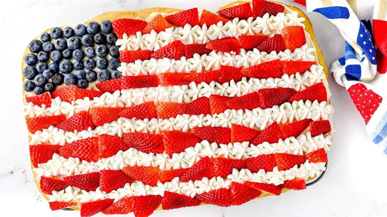 An easy American Flag Cookie Cake decorated with blueberries, strawberries, and whipped cream, perfect as a 4th of July dessert on a white surface with a colorful cloth nearby.