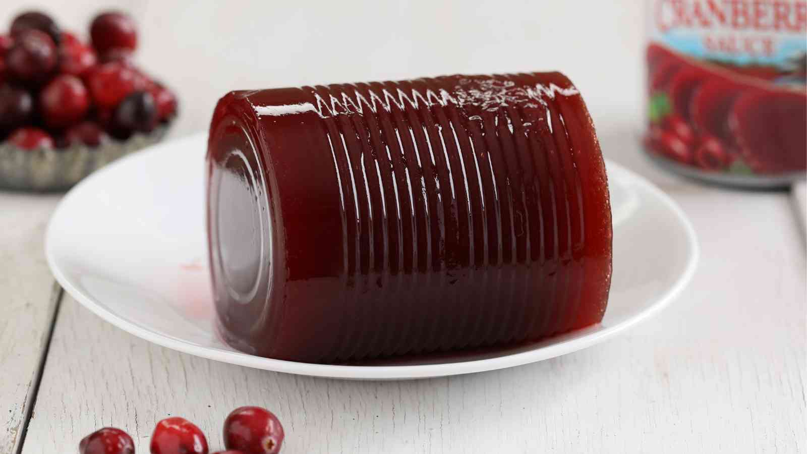 A cylinder-shaped block of canned cranberry sauce sits on a white plate with whole cranberries nearby and part of a can visible in the background.