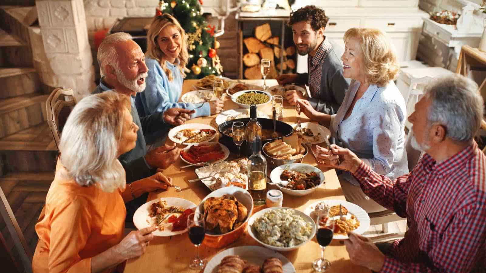 Six adults sit around a dining table sharing a festive meal, with various dishes and drinks, and a decorated Christmas tree in the background.