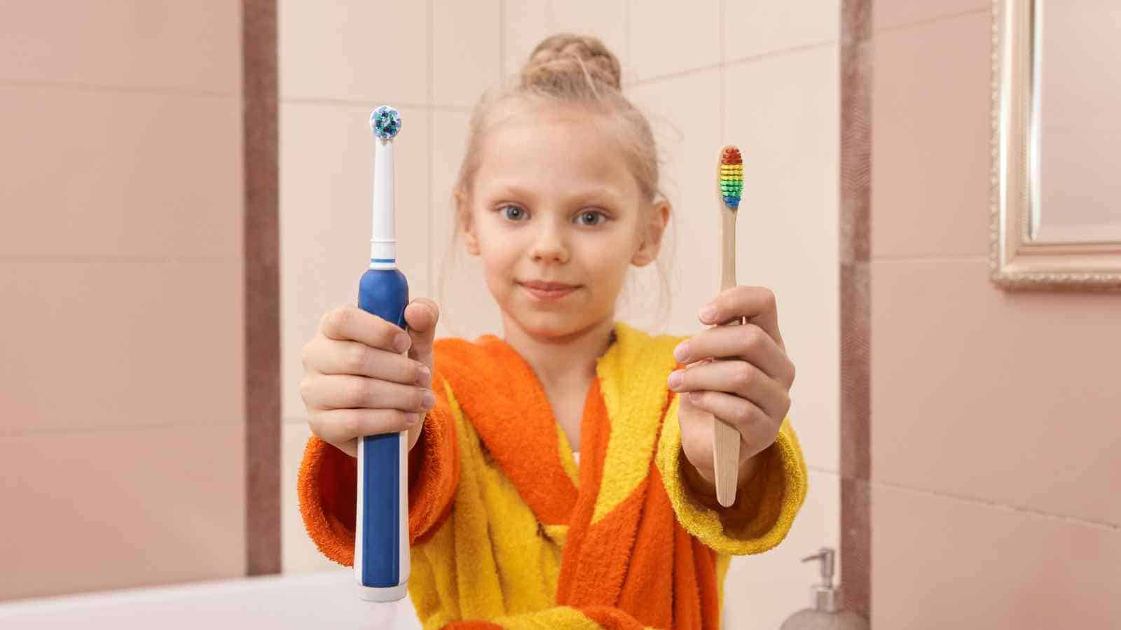 A child in a yellow and orange robe holds an electric toothbrush in one hand and a manual toothbrush in the other in a bathroom.