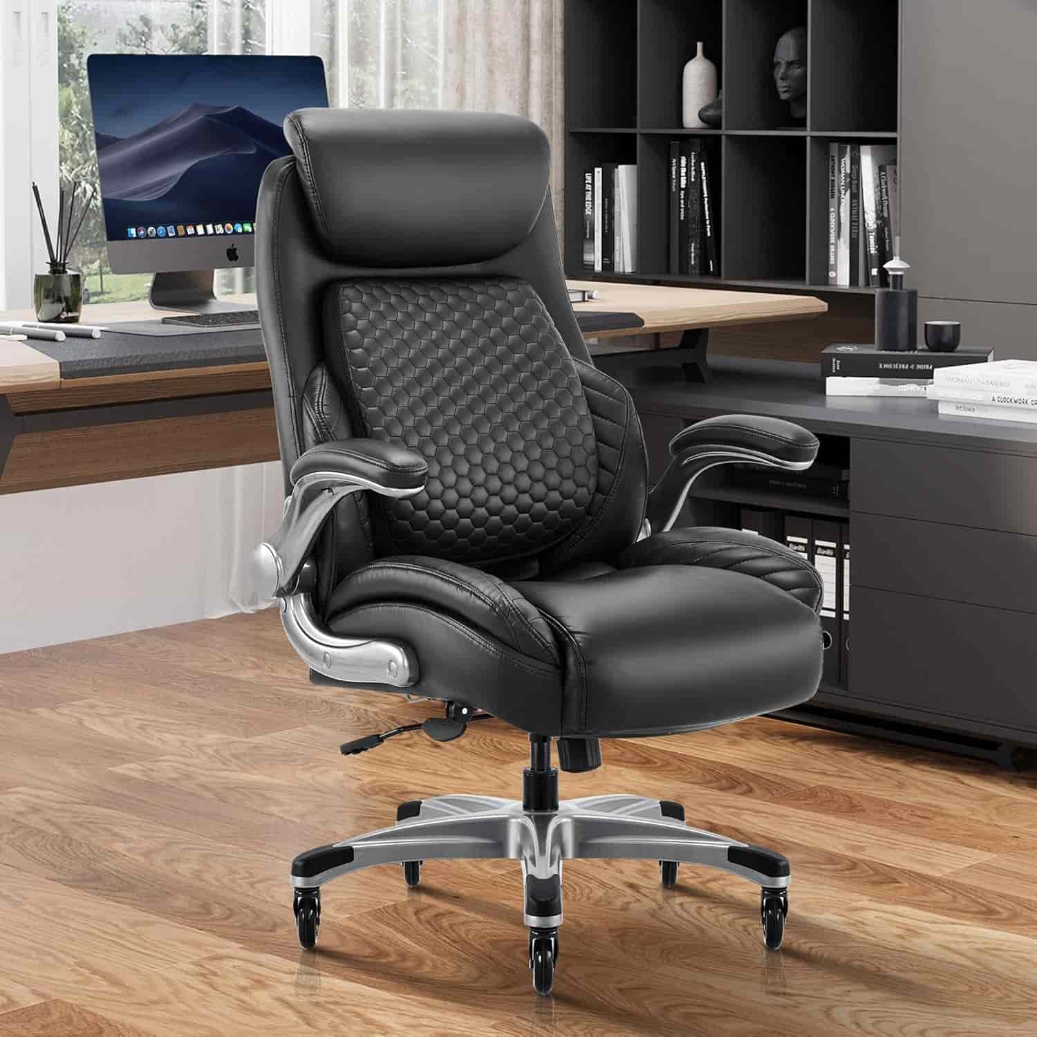 A black ergonomic office chair with padded seat and backrest on wheels sits in a modern office with a desk, computer, and shelves in the background.