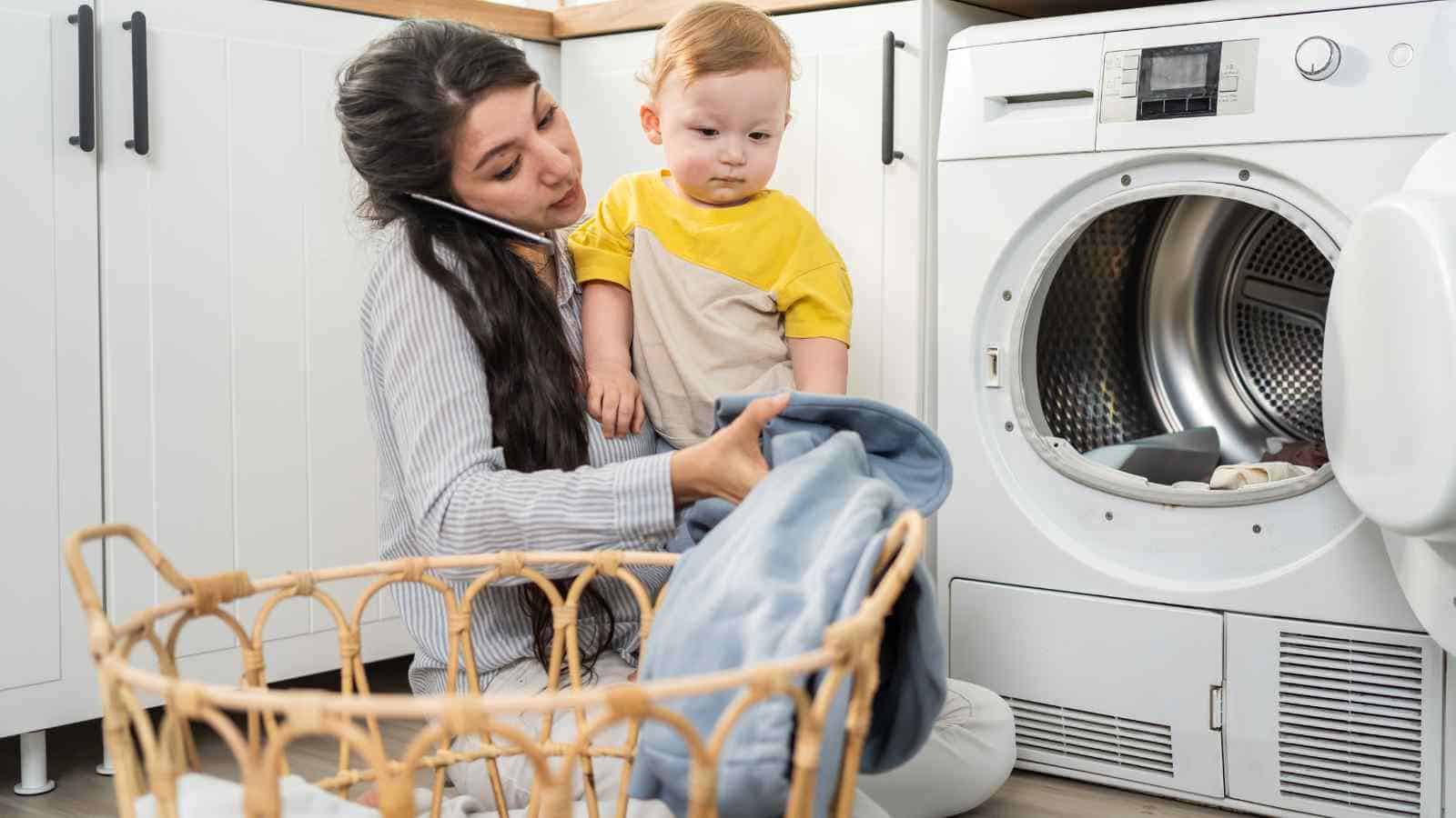 A woman multitasks by talking on the phone while holding a toddler and loading laundry into a front-loading washing machine in a laundry room.