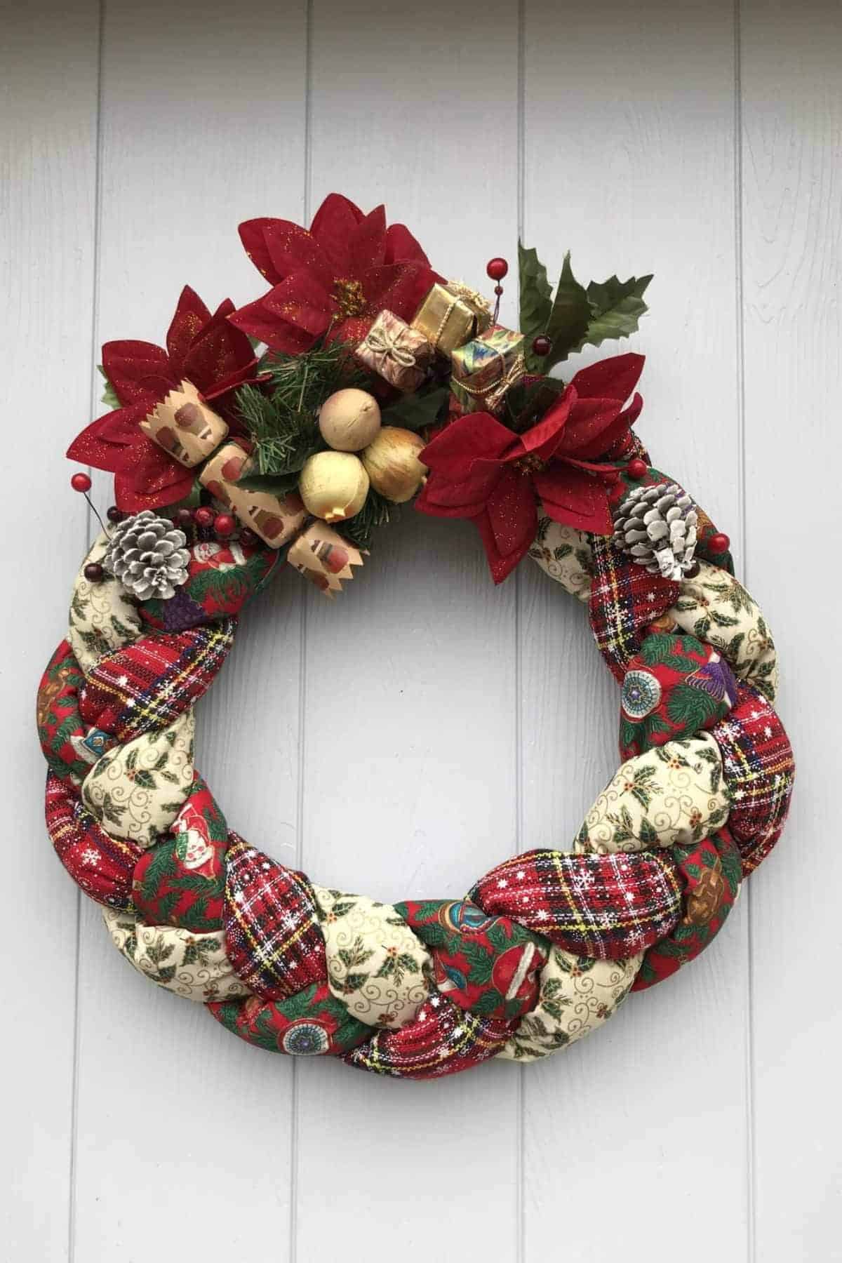 A braided Christmas wreath decorated with red poinsettias, pinecones, holly leaves, small presents, and artificial apples, hanging on a light-colored wall.