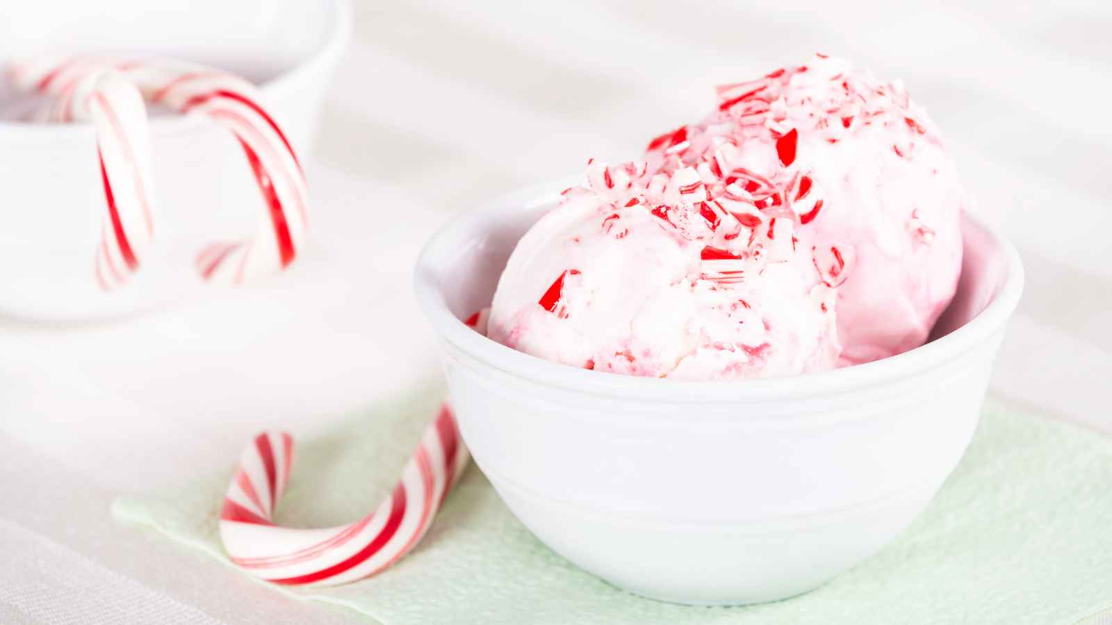 Two scoops of ice cream topped with crushed candy canes in a white bowl, with candy canes and another bowl in the background.