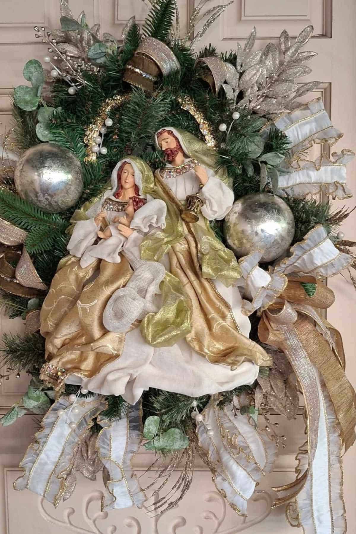 A Christmas wreath decorated with metallic ornaments, ribbons, greenery, and figurines of Mary, Joseph, and baby Jesus in the center.