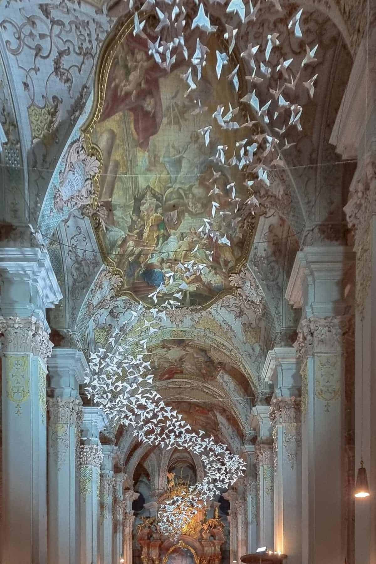 A large group of white origami birds is suspended from the ornate, vaulted ceiling of a church with detailed murals and classical columns.