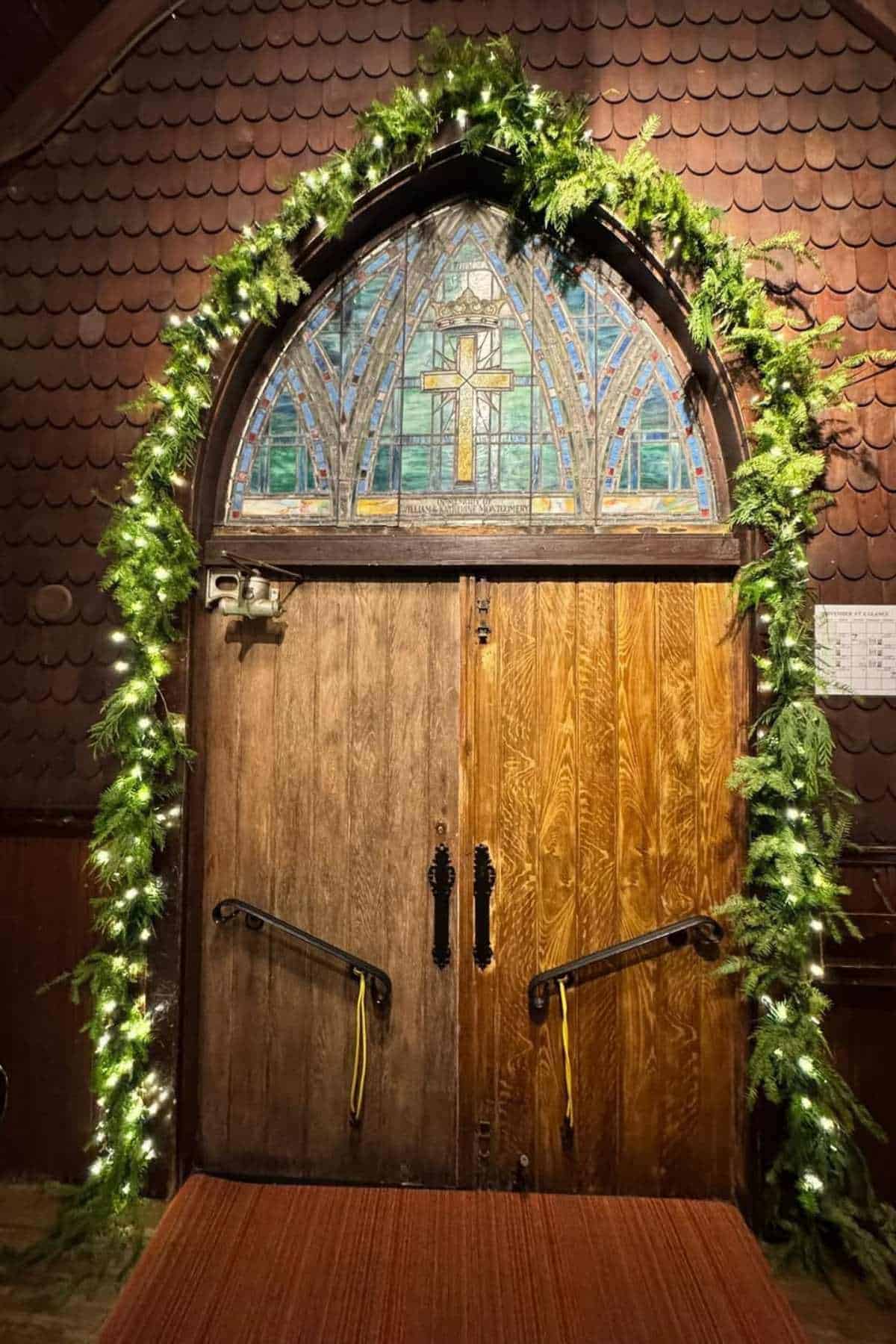 Wooden double doors with iron handles, framed by greenery and string lights, beneath a stained glass window featuring a cross.