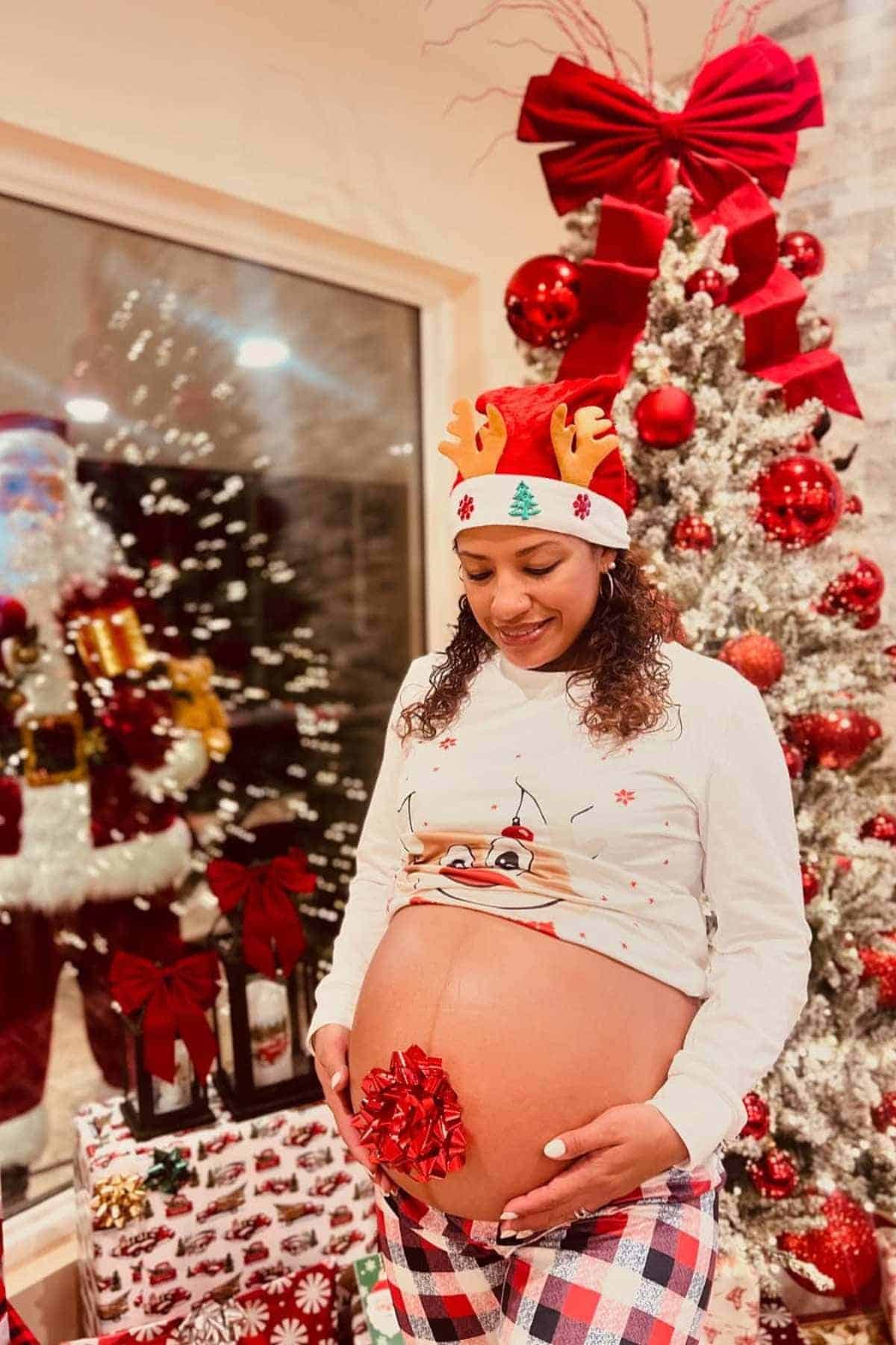 A pregnant woman wearing a festive outfit stands in front of a decorated Christmas tree, holding her belly with a red bow on it and presents in the background.