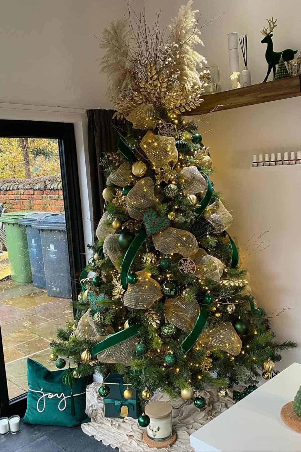 A decorated Christmas tree with gold and green ornaments, ribbon, and lights stands in a living room near a window and green pillows.