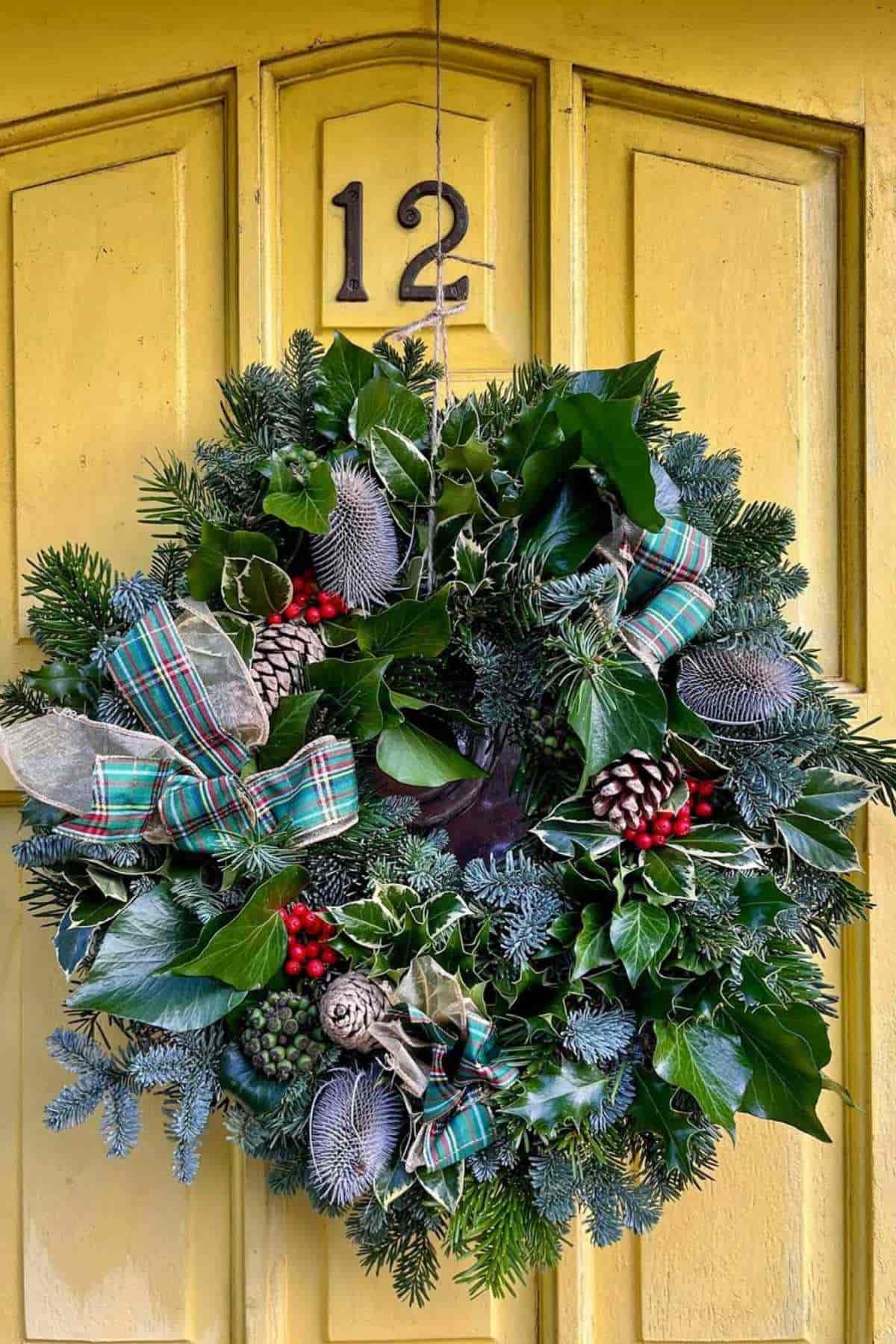 A festive wreath with green foliage, pinecones, red berries, and plaid ribbon hangs on a yellow door with the number 12 above it.