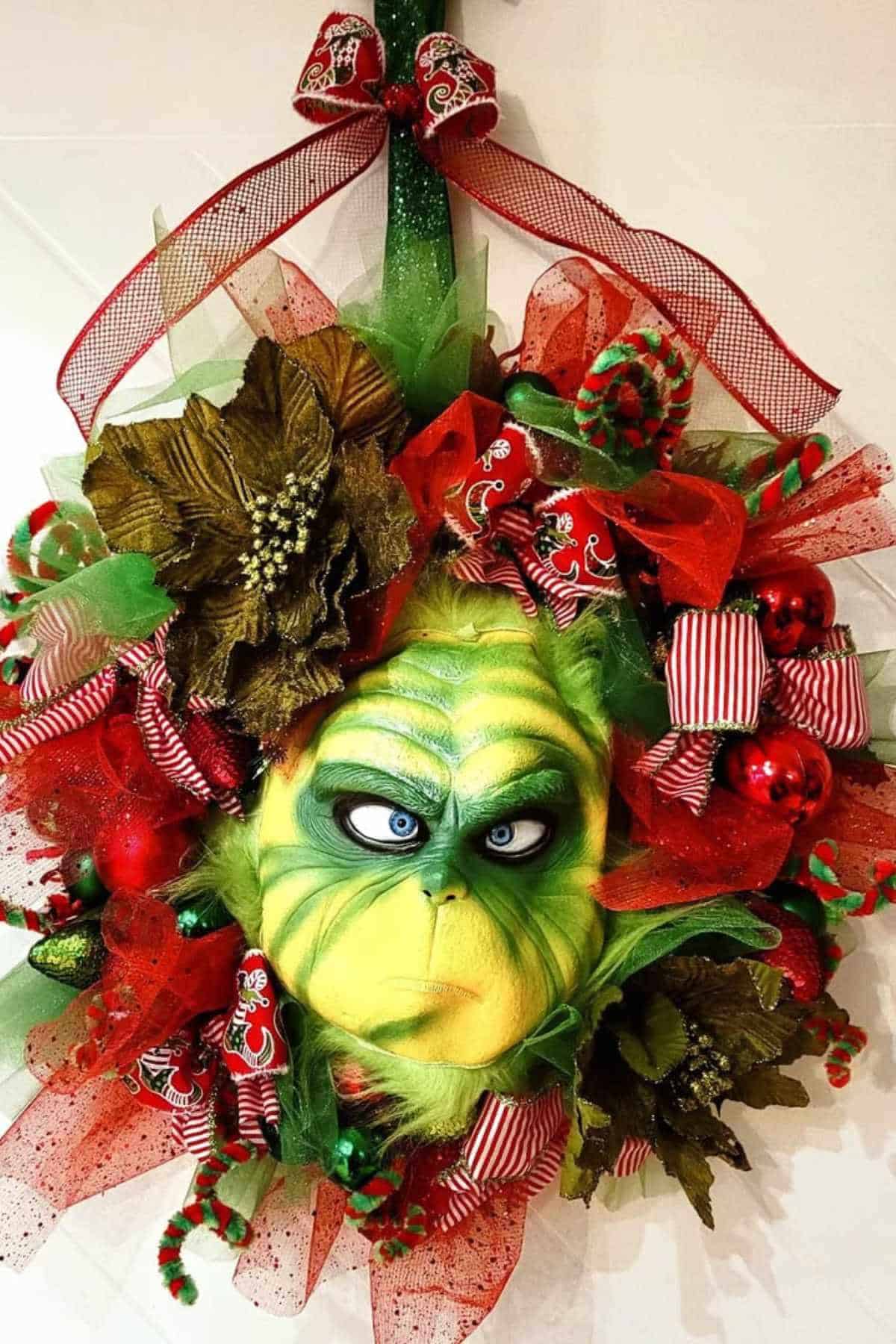 A Christmas wreath decorated with red, green, and gold ornaments, featuring a central mask of the Grinch with a red bow on top.