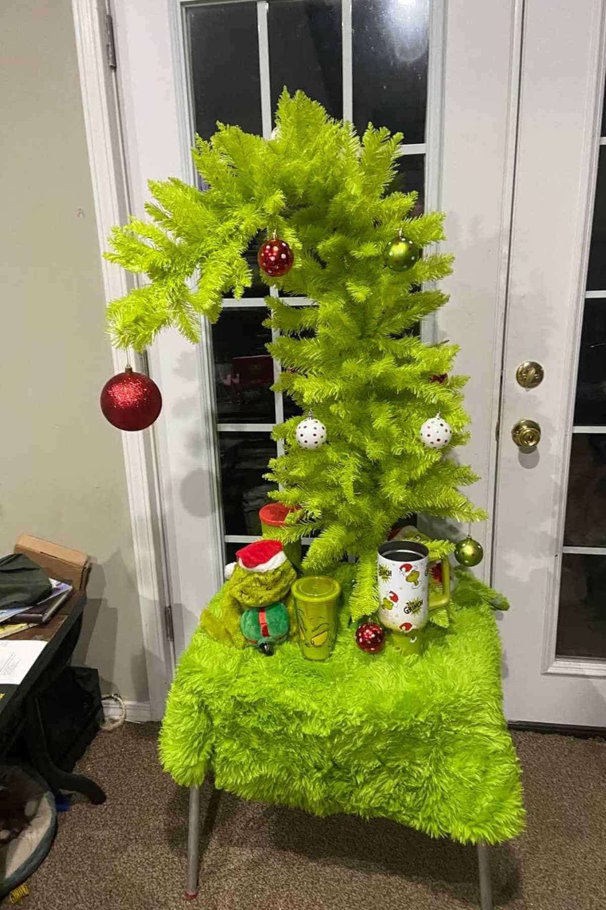 A small, bright green artificial Christmas tree with a curved top is decorated with ornaments and placed on a matching green table in front of glass doors.