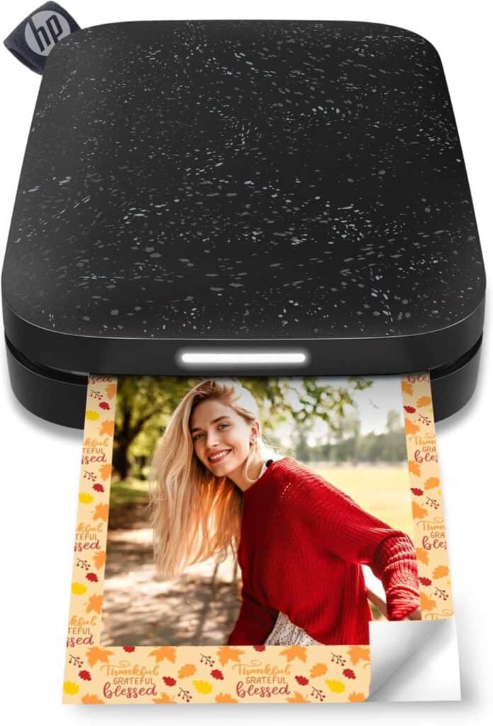 A compact HP photo printer prints a color photograph of a smiling woman wearing a red sweater, with a festive autumn border.
