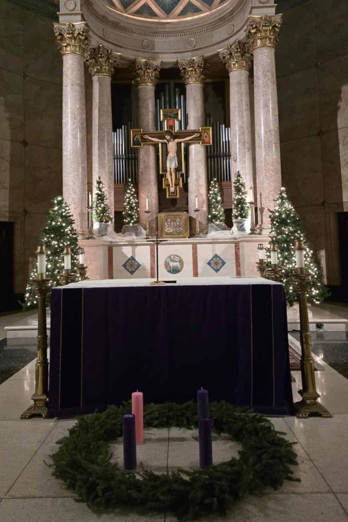 An altar decorated with lit Christmas trees, a crucifix, and a purple cloth, with four Advent candles in a wreath in the foreground.