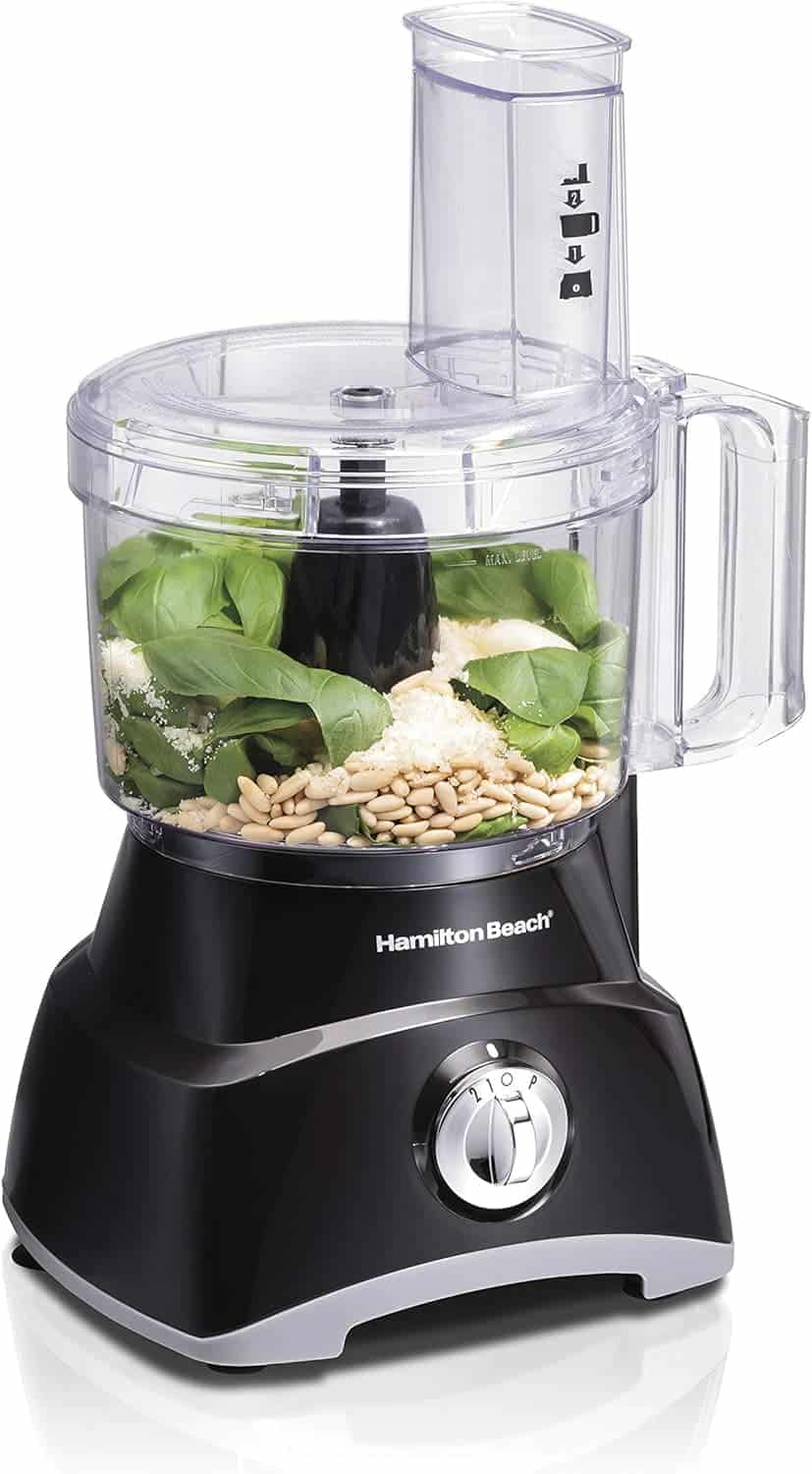 A Hamilton Beach food processor with basil leaves, pine nuts, and grated cheese inside the bowl, ready for processing.
