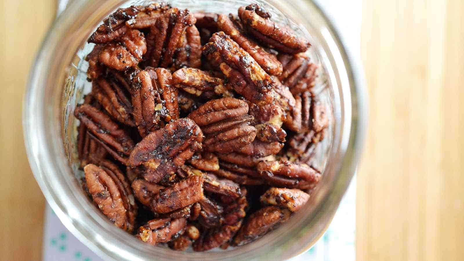 A glass jar filled with roasted pecans viewed from above on a light-colored surface.