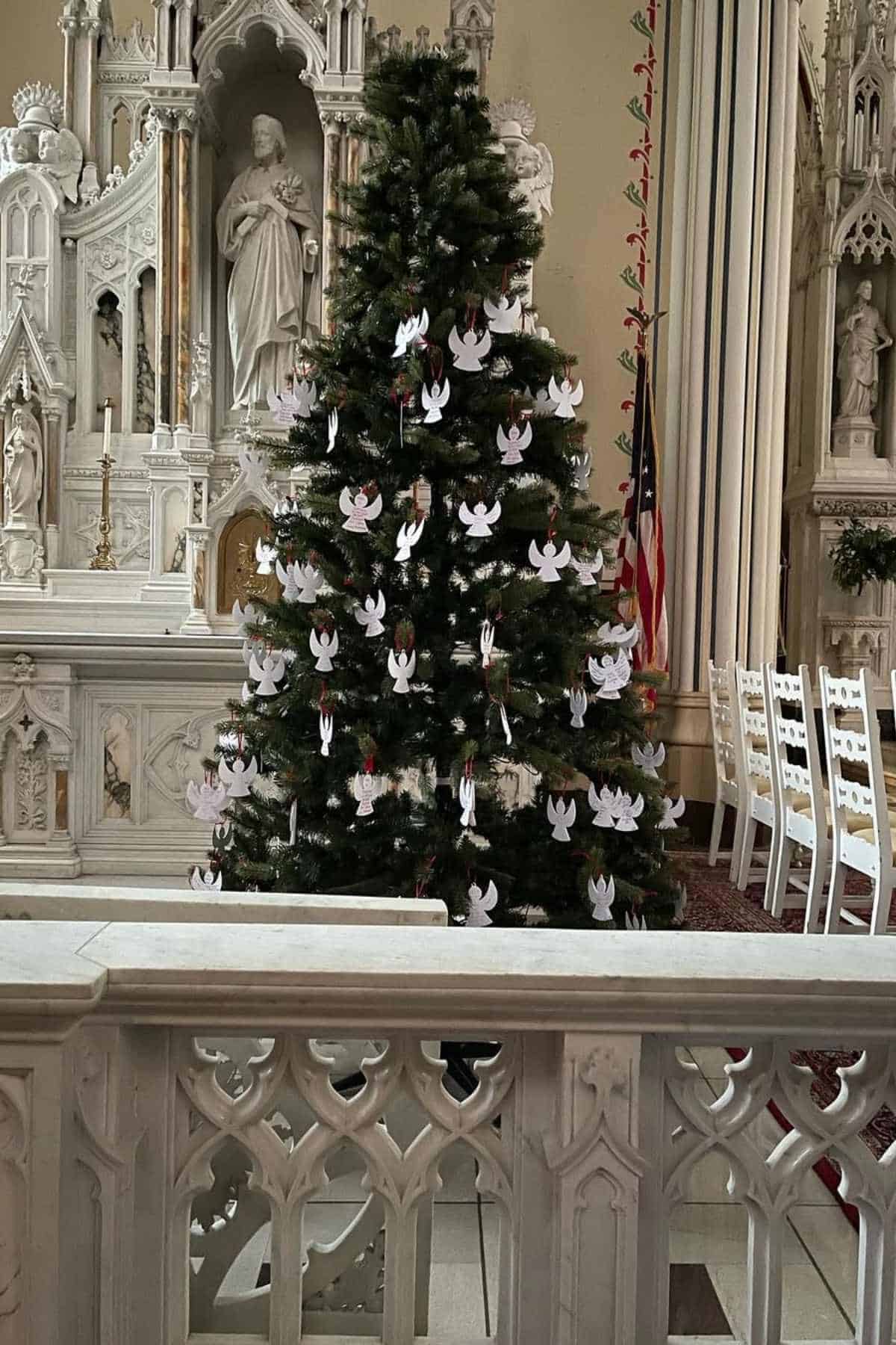 A decorated Christmas tree with white paper angel stands inside a church, surrounded by ornate white altars and statues.