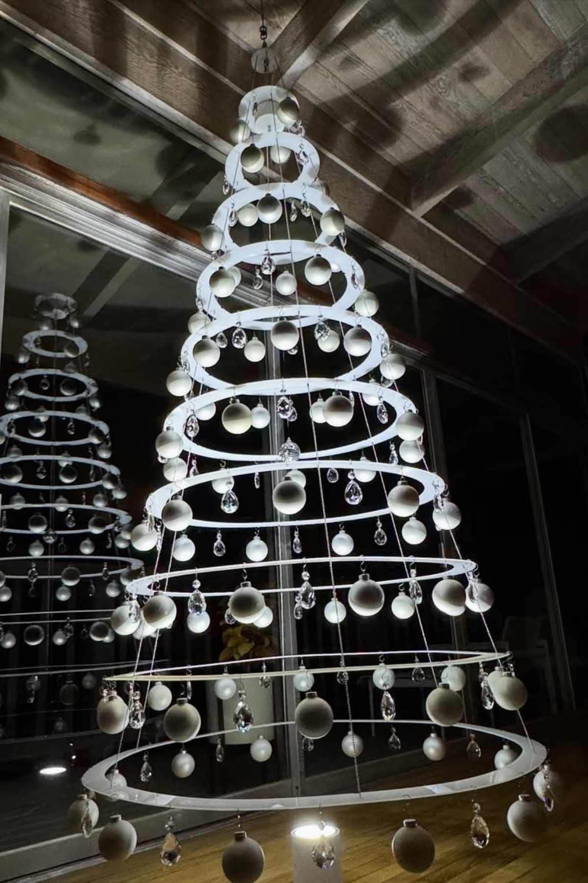A modern Christmas tree made of suspended white rings, decorated with white ornaments and crystal drops, stands indoors with its reflection visible in a glass window behind.