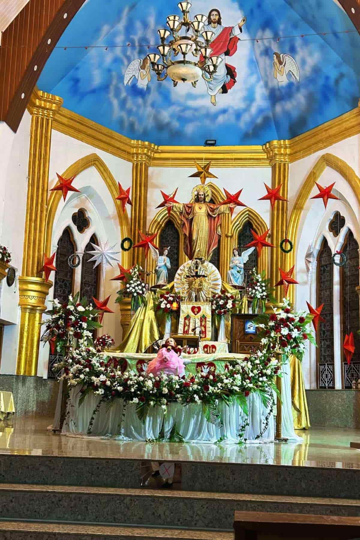A church altar decorated with red stars, flowers, and religious statues, with a painted ceiling showing Jesus and angels.