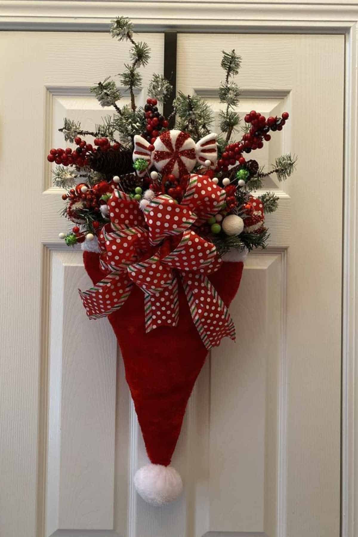 A red Santa hat-shaped door hanging filled with artificial greenery, red berries, candy decorations, and a large red polka-dot bow.