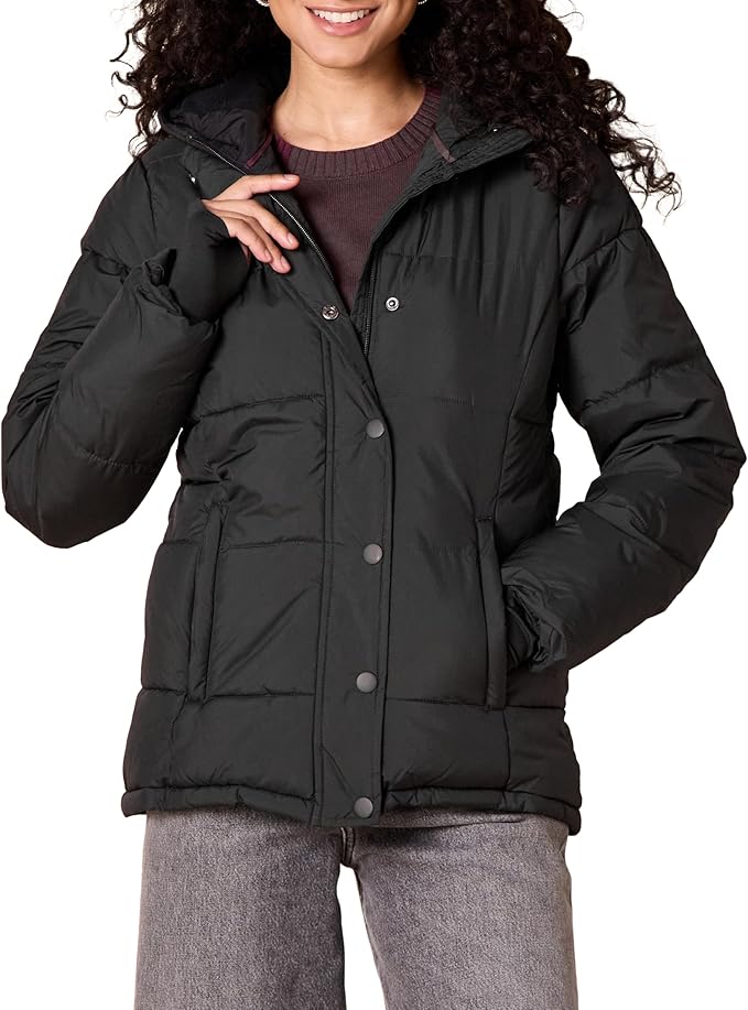 A person wearing a black puffer jacket over a brown sweater and gray pants, standing with one hand touching the jacket’s collar.