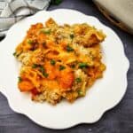 A serving of baked pasta with sauce, melted cheese, and parsley on a white plate, placed on a dark surface next to a checkered cloth.