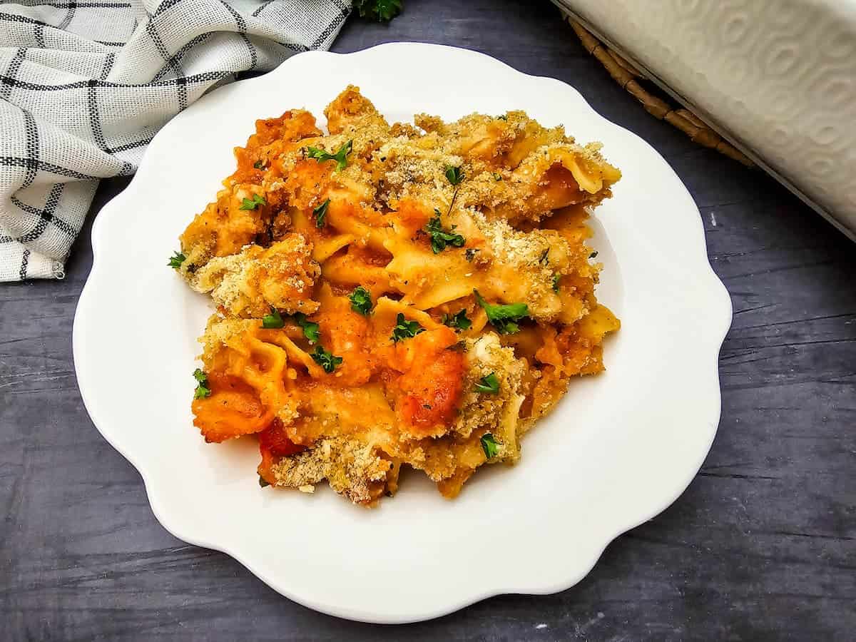 A serving of baked pasta with sauce, melted cheese, and parsley on a white plate, placed on a dark surface next to a checkered cloth.