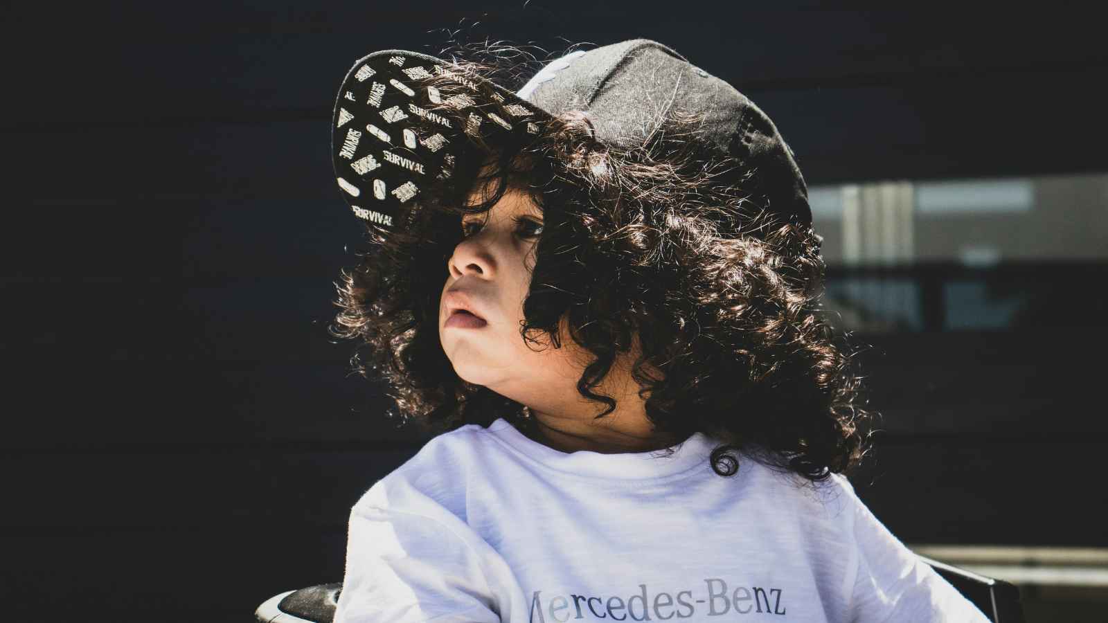 Young child with curly hair wears a black cap sideways and a white Mercedes-Benz T-shirt, looking off to the side against a dark background.