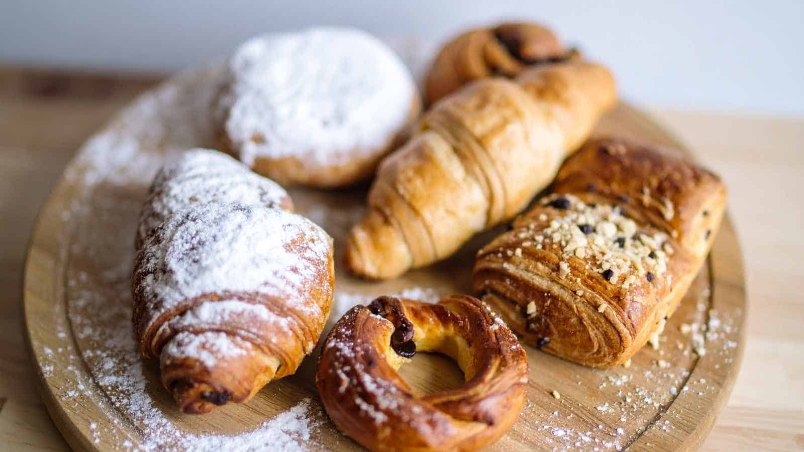 A round wooden board with six assorted pastries, including croissants, danishes, and pastries dusted with powdered sugar.