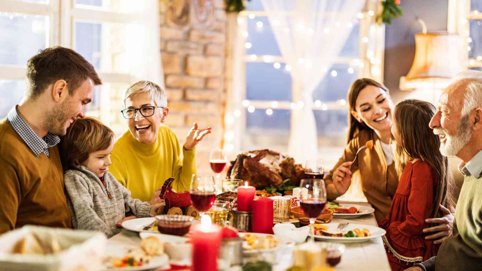 Six people of varying ages sit around a dinner table with food, drinks, and candles, smiling and engaging in conversation in a warmly lit room.