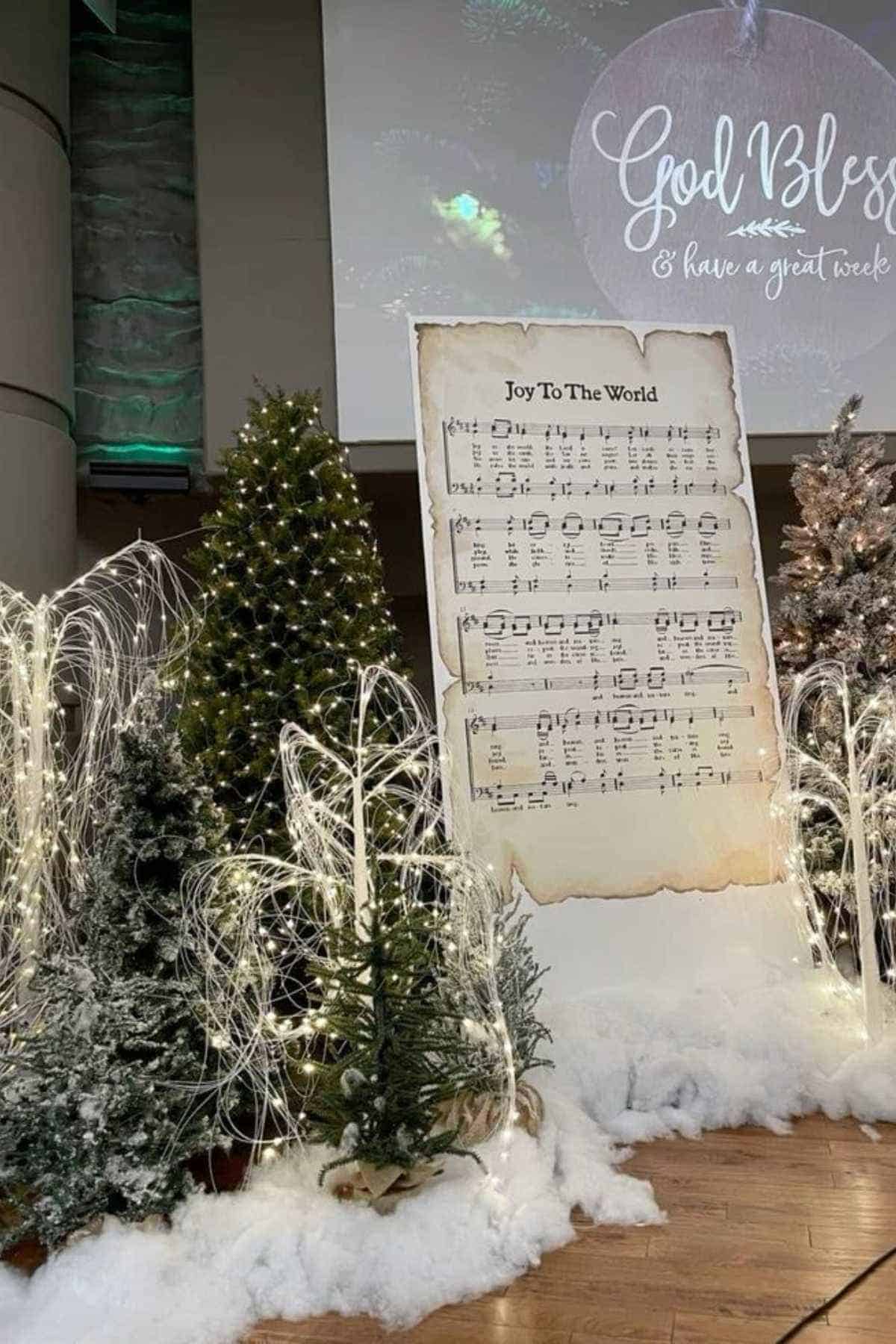 Christmas-themed stage display with decorated trees, artificial snow, string lights, and a large sheet of "Joy To The World" music. A "God Bless" projection is visible in the background.
