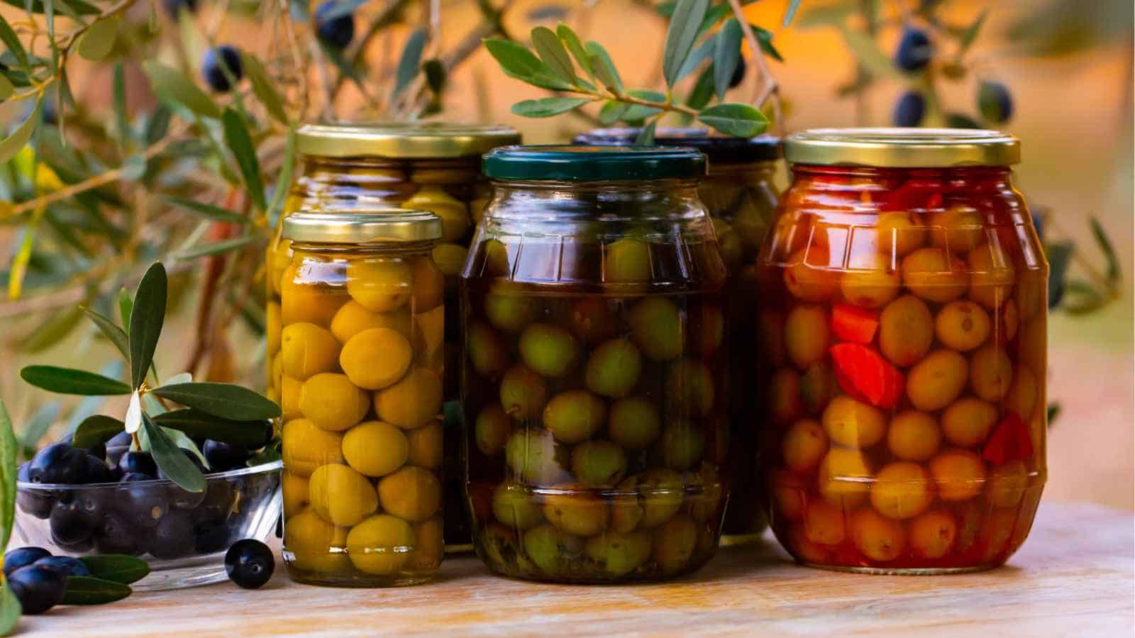 Five jars of assorted olives preserved in liquid, with a small glass bowl of fresh olives and olive branches in the background.