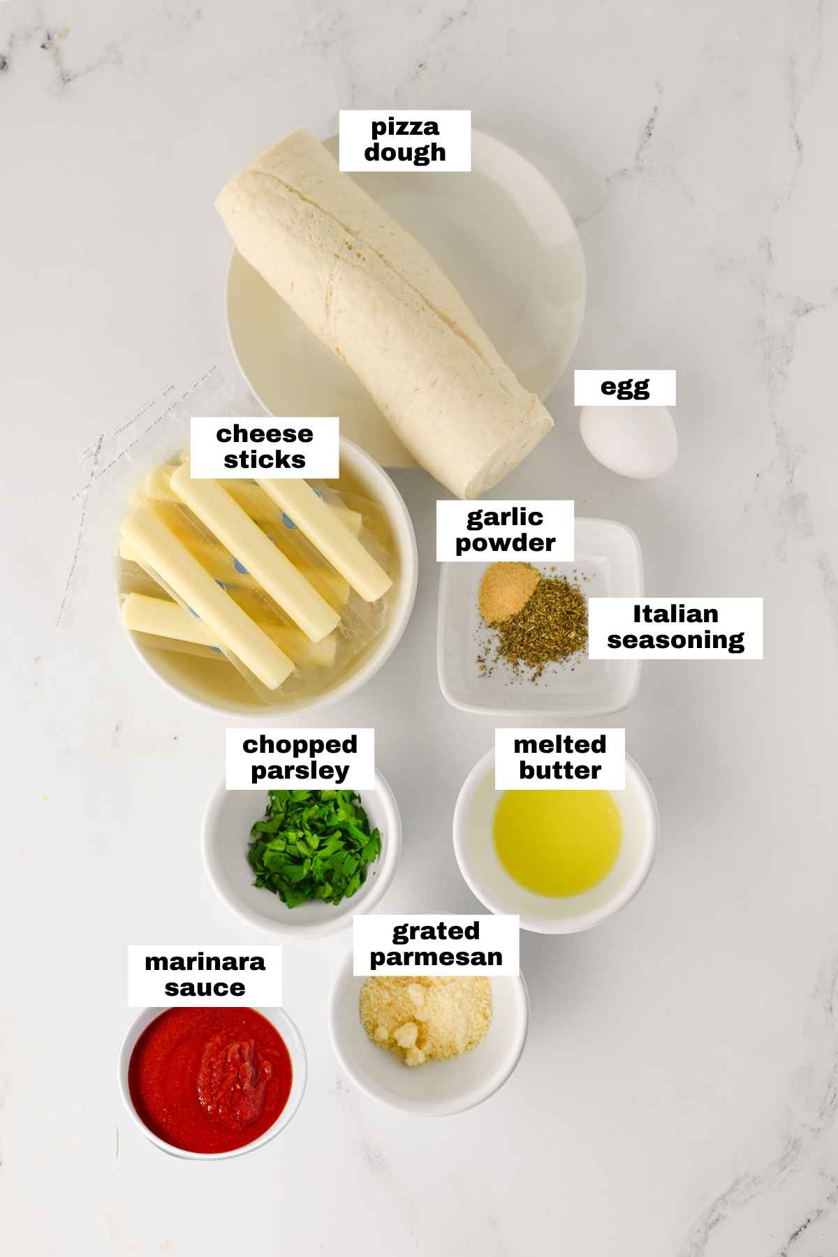 Ingredients for a recipe displayed on a white surface: pizza dough, cheese sticks, egg, garlic powder, Italian seasoning, chopped parsley, melted butter, grated parmesan, and marinara sauce.