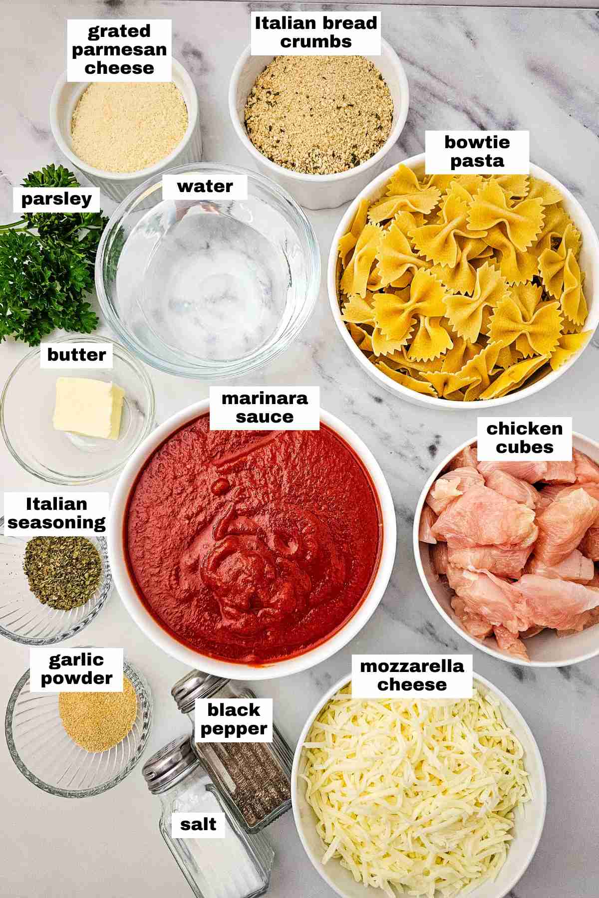 Labeled ingredients for a pasta dish arranged on a marble surface, including bowtie pasta, chicken cubes, marinara sauce, cheeses, bread crumbs, butter, seasonings, parsley, and water.
