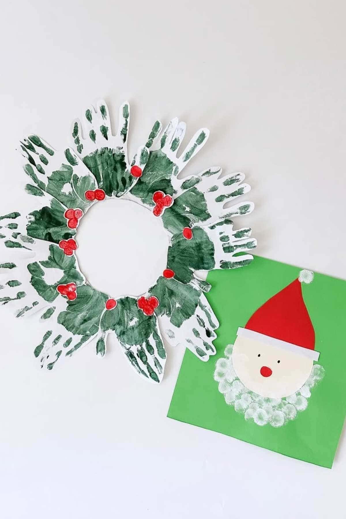 A Christmas wreath made of green handprints with red berries, next to a Santa face craft with a red hat and a beard made from white paint dots on green paper.
