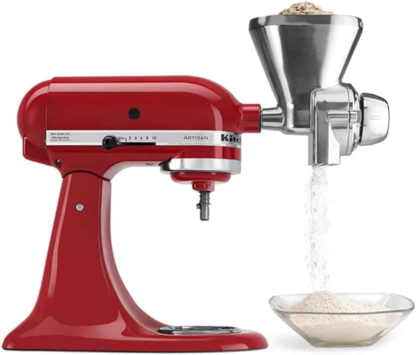 A red stand mixer with a metal grain mill attachment is grinding flour into a glass bowl.