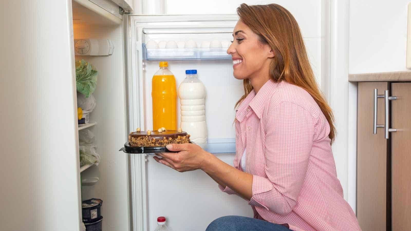 A woman kneels in front of an open refrigerator, smiling as she holds a chocolate cake. There are bottles of juice and milk on the refrigerator door shelves.