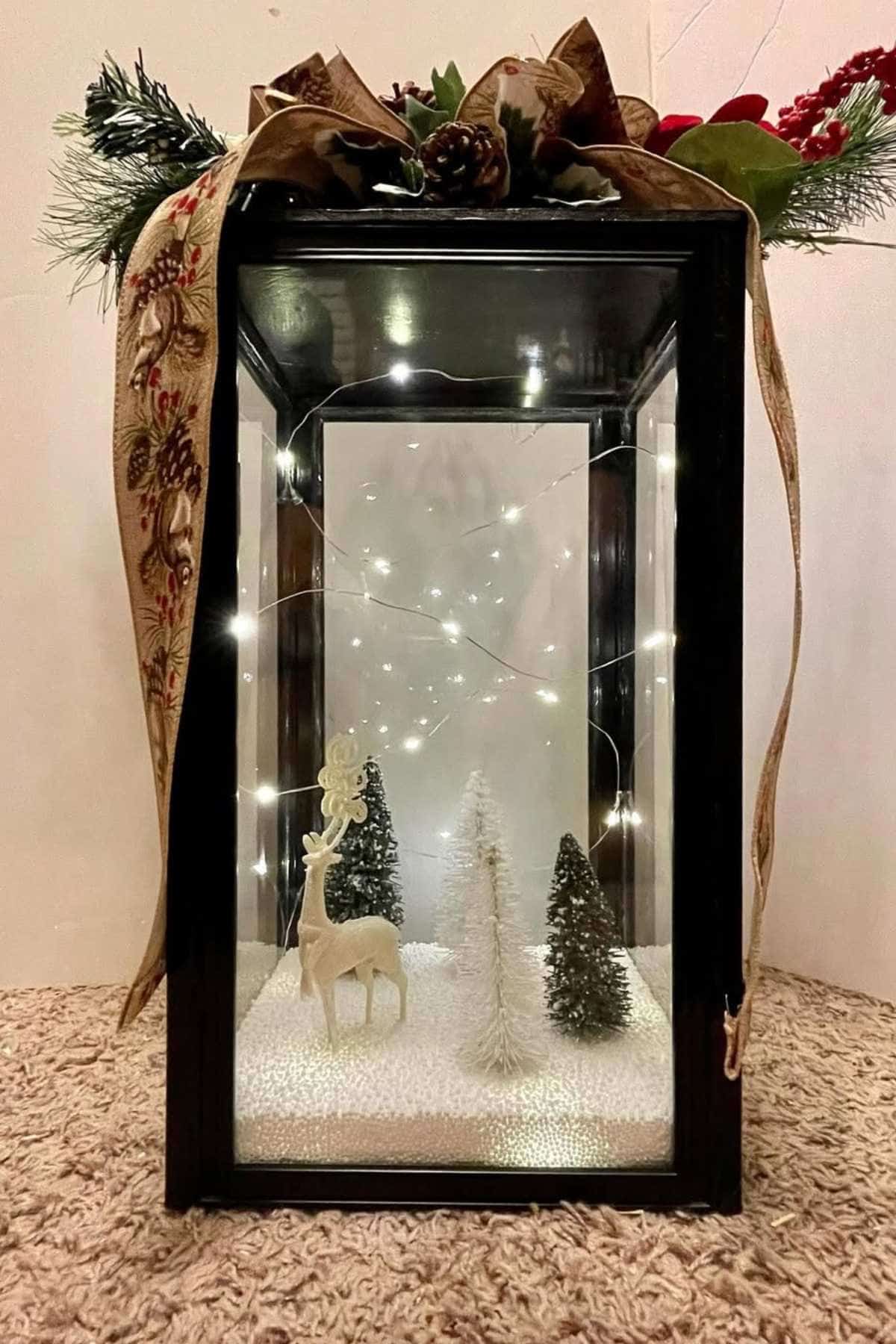 A black-framed glass box displays a white reindeer and three trees on artificial snow, with string lights and a decorative ribbon and greenery on top.