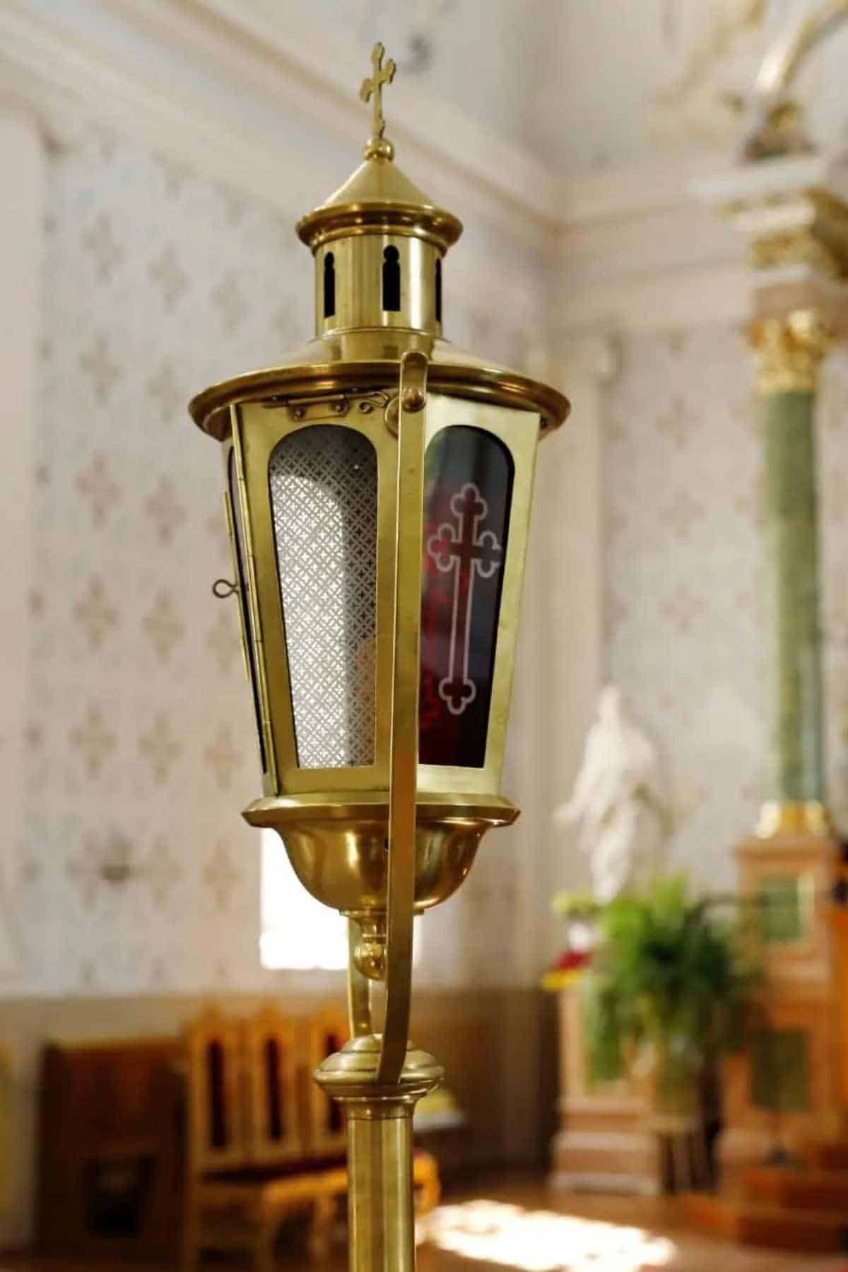 A brass sanctuary lamp with red and clear glass panels, featuring a cross design, stands inside a church with ornate decor in the background.