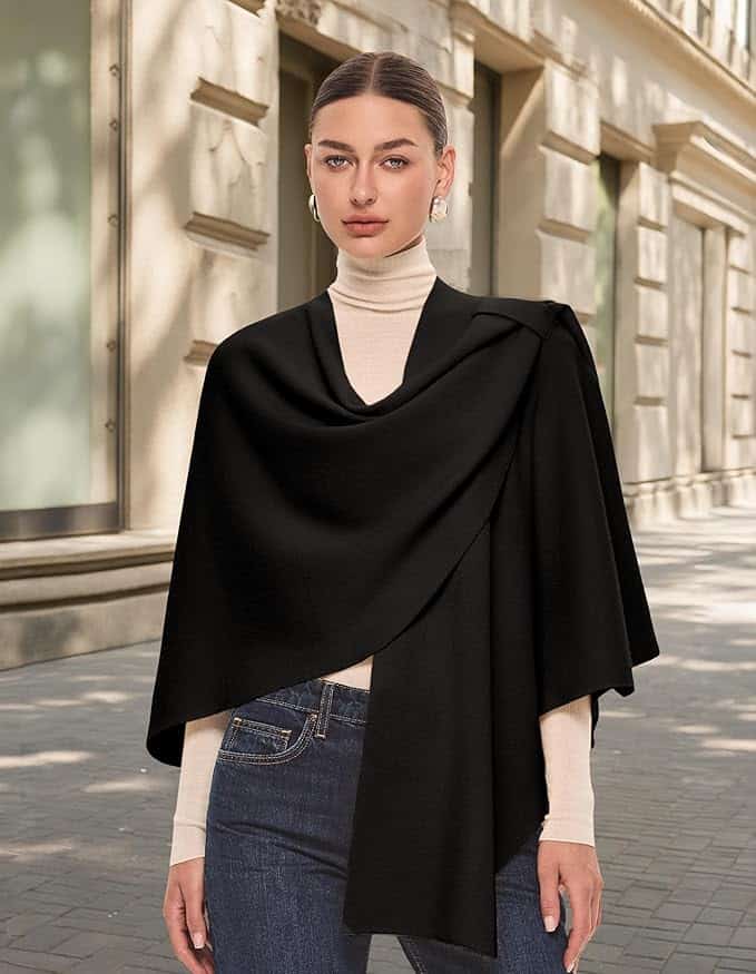 Woman standing on a city sidewalk, wearing a cream turtleneck, blue jeans, and a black asymmetrical cape.