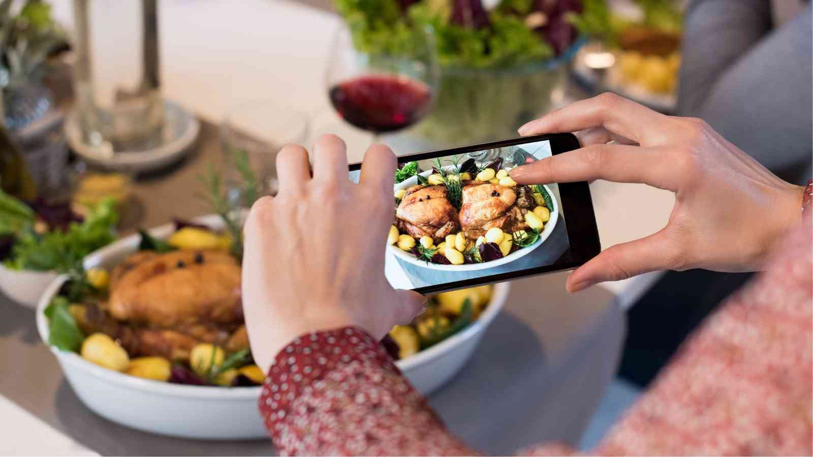 A person takes a photo of a roasted chicken dish with vegetables using a smartphone at a dining table.