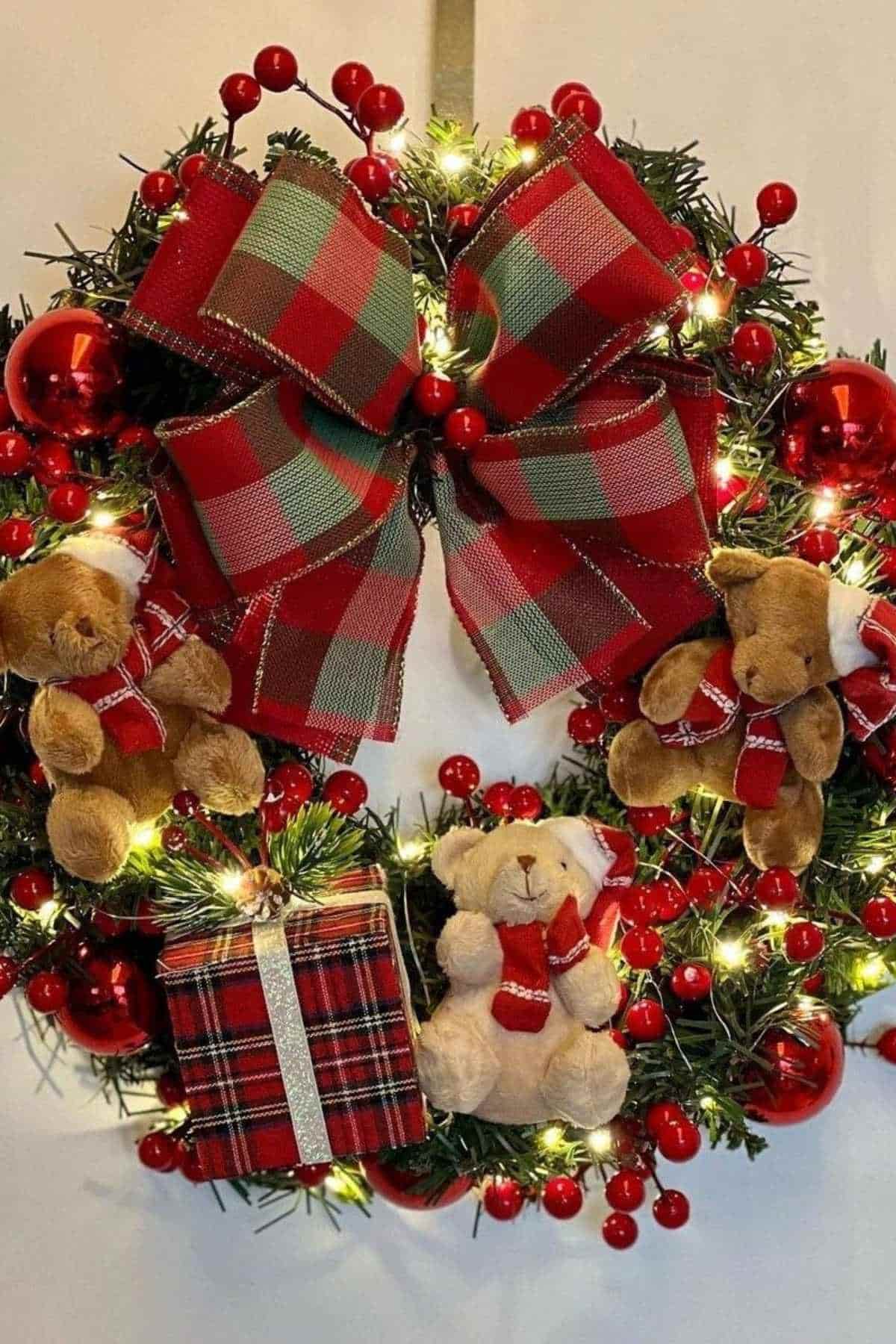 A festive Christmas wreath decorated with red plaid ribbon, red ornaments, small teddy bears, red berries, and a plaid-wrapped gift, all accented with string lights.