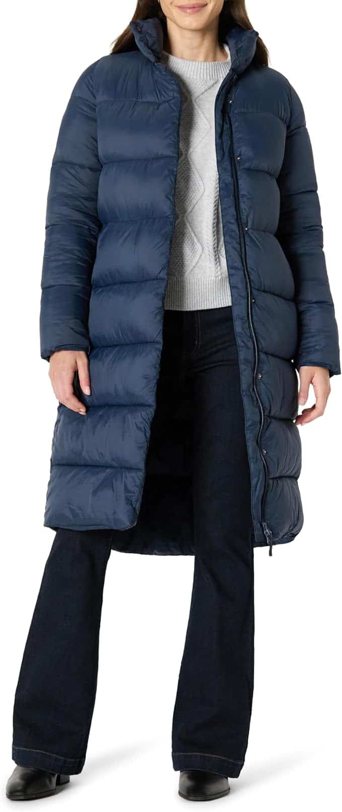 A person wearing a long, blue puffer coat over a light gray sweater, dark jeans, and black shoes, standing against a plain white background.