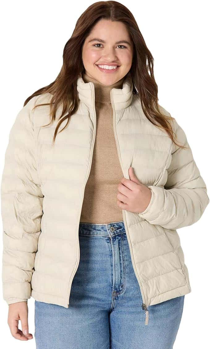 A woman wearing a beige puffer jacket over a tan sweater and blue jeans stands smiling against a plain background.