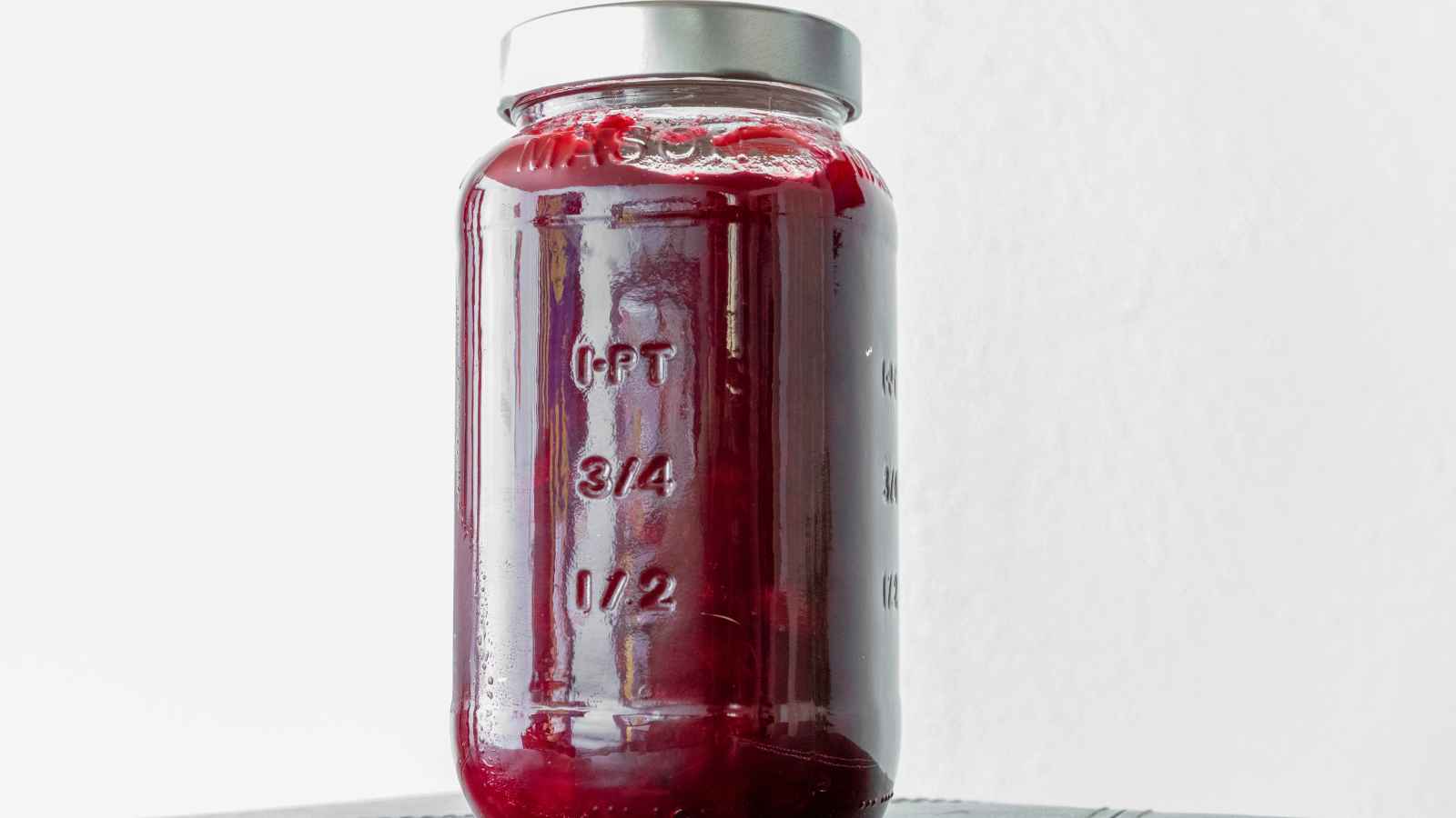 A glass jar filled with red jam or preserves, sealed with a metal lid. Measurement markings are visible on the side of the jar.