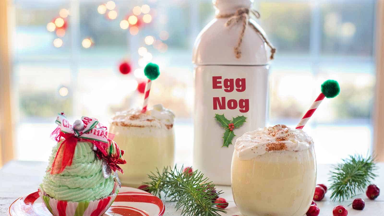 A bottle labeled "Egg Nog" with two glasses of eggnog, topped with whipped cream and straws, beside a green cupcake decorated with holiday ribbons.
