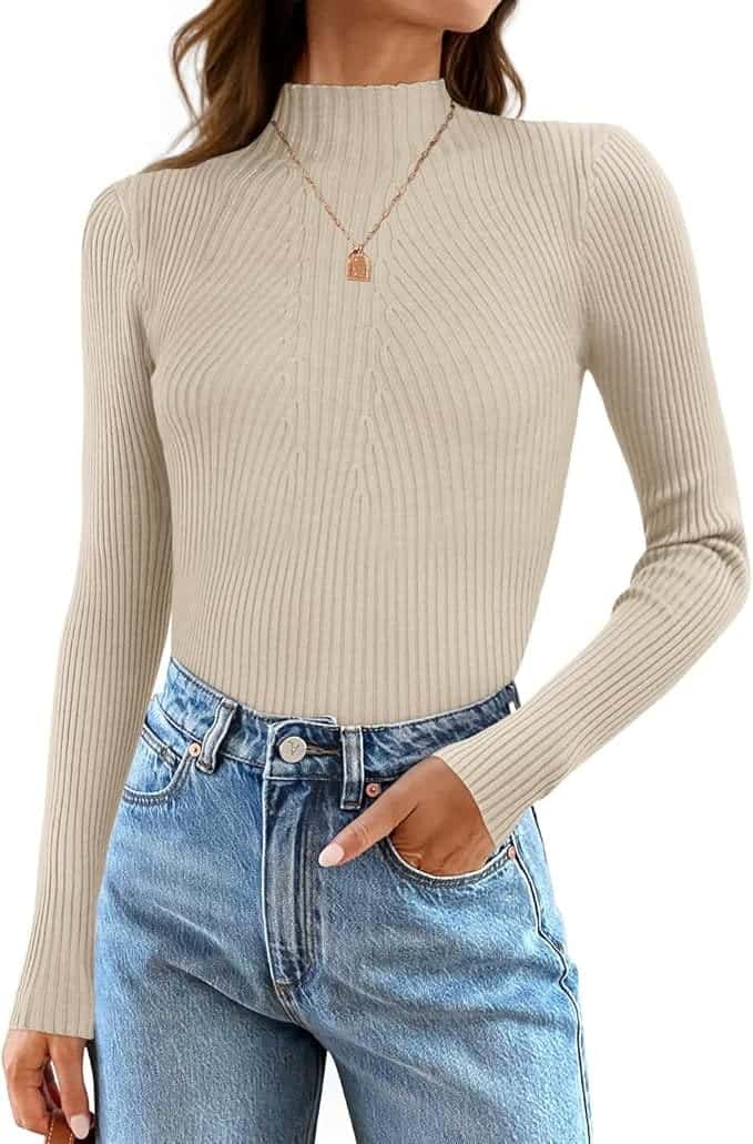 A person wearing a beige ribbed long-sleeve turtleneck sweater, blue jeans, and a gold necklace with a square pendant.