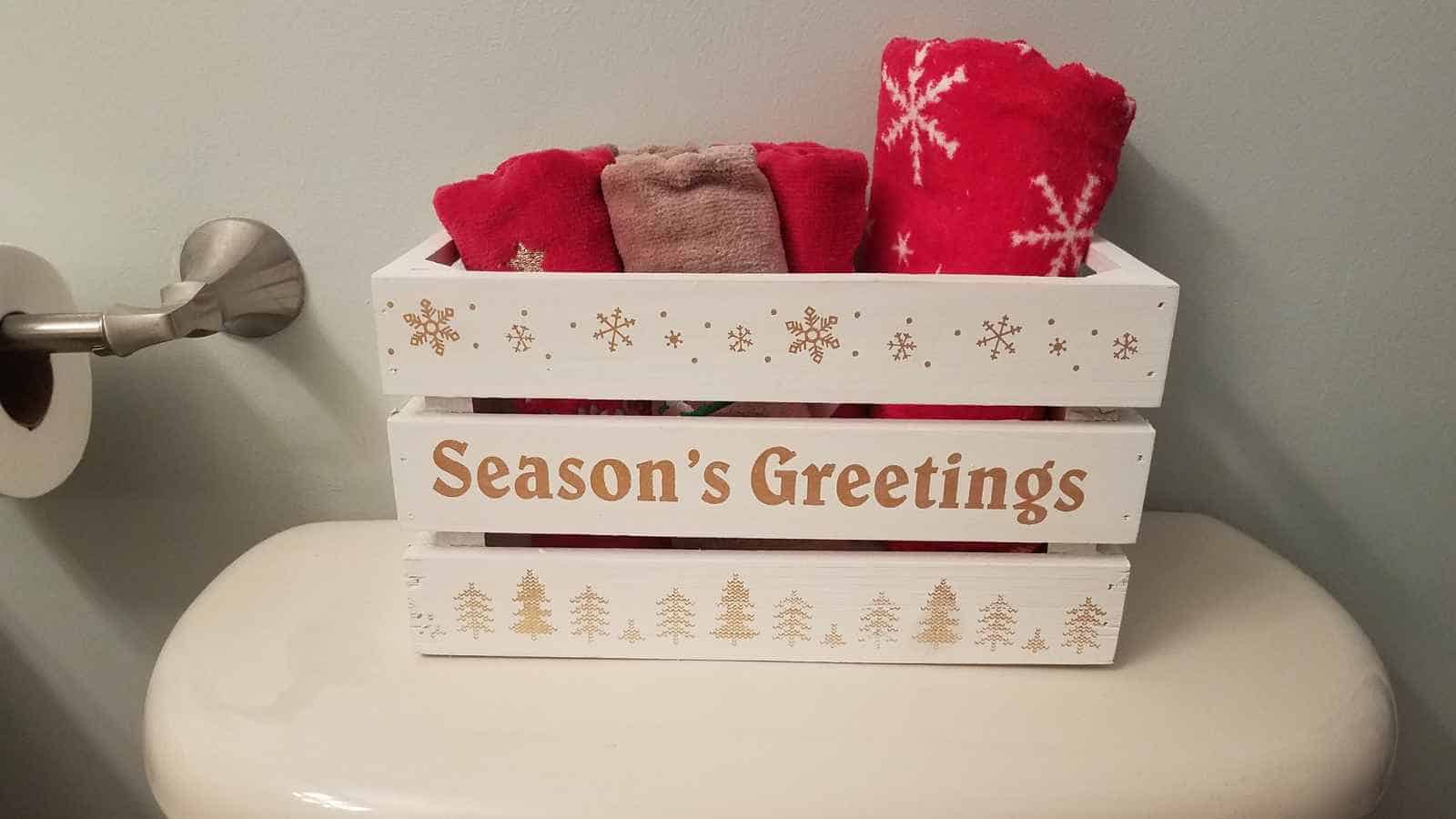 A white wooden box labeled "Season's Greetings" holds red and beige towels and is placed on top of a toilet tank next to a toilet paper holder.